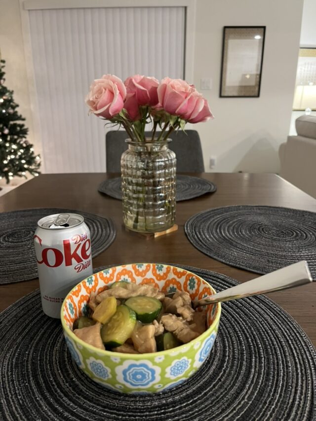 picture of a bowl of food filled with chicken and zucchini, a can of diet coke, and vase of pink roses on a wooden dining room table. 